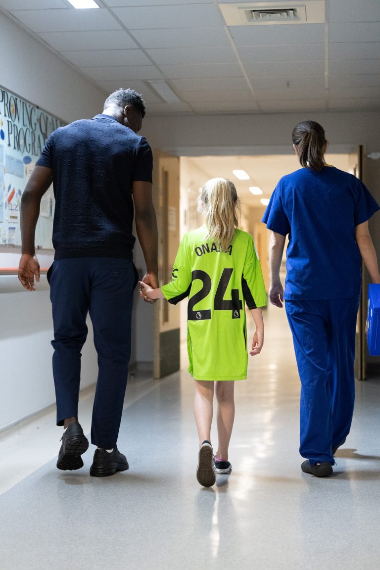 Andre Onana walks with young girl wearing special Onana hospital gowns and nurse down hospital corridor