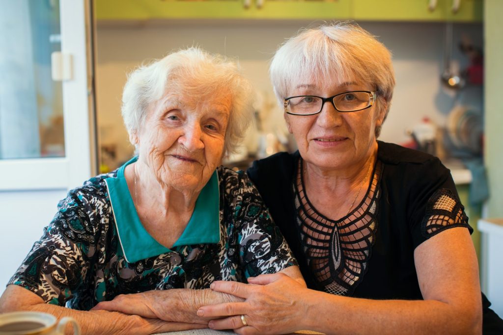 Two older women sit next to each other arm in arm in a hospital environment.